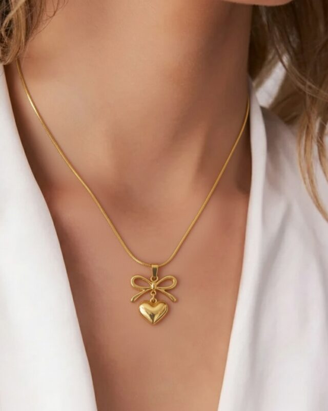 A delicate gold heart-shaped pendant with a tied ribbon design, placed on a soft neutral background. The pendant’s polished surface glows under light, showcasing its romantic and elegant craftsmanship.