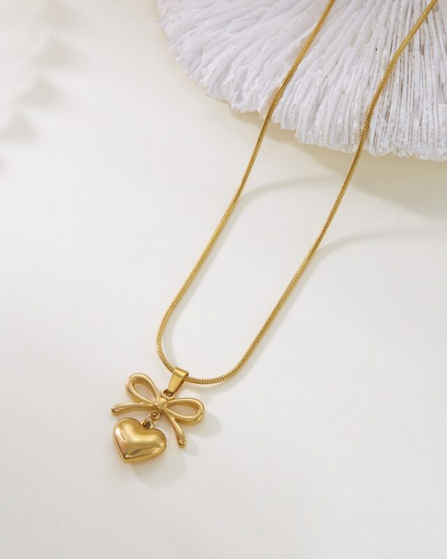 A delicate gold heart-shaped pendant with a tied ribbon design, placed on a soft neutral background. The pendant’s polished surface glows under light, showcasing its romantic and elegant craftsmanship.